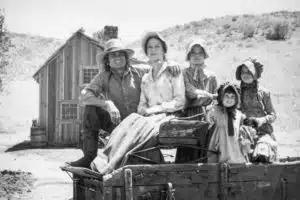 A family of five in pioneer-era clothing poses outdoors by a wooden house. The parents and three children, all dressed in bonnets and hats, sit on a wooden cart. The landscape is open and sparse, much like the dedication you’ll find with Georgia Probate Law Group - Your Professional Probate Attorney.