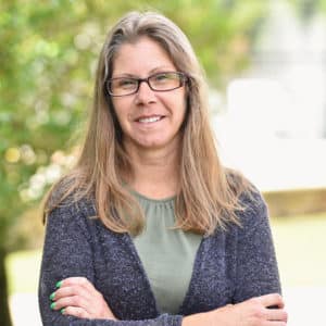 Matisha Pittman, a woman with long hair and glasses, wearing a green shirt and gray cardigan, stands outdoors with arms crossed, smiling.